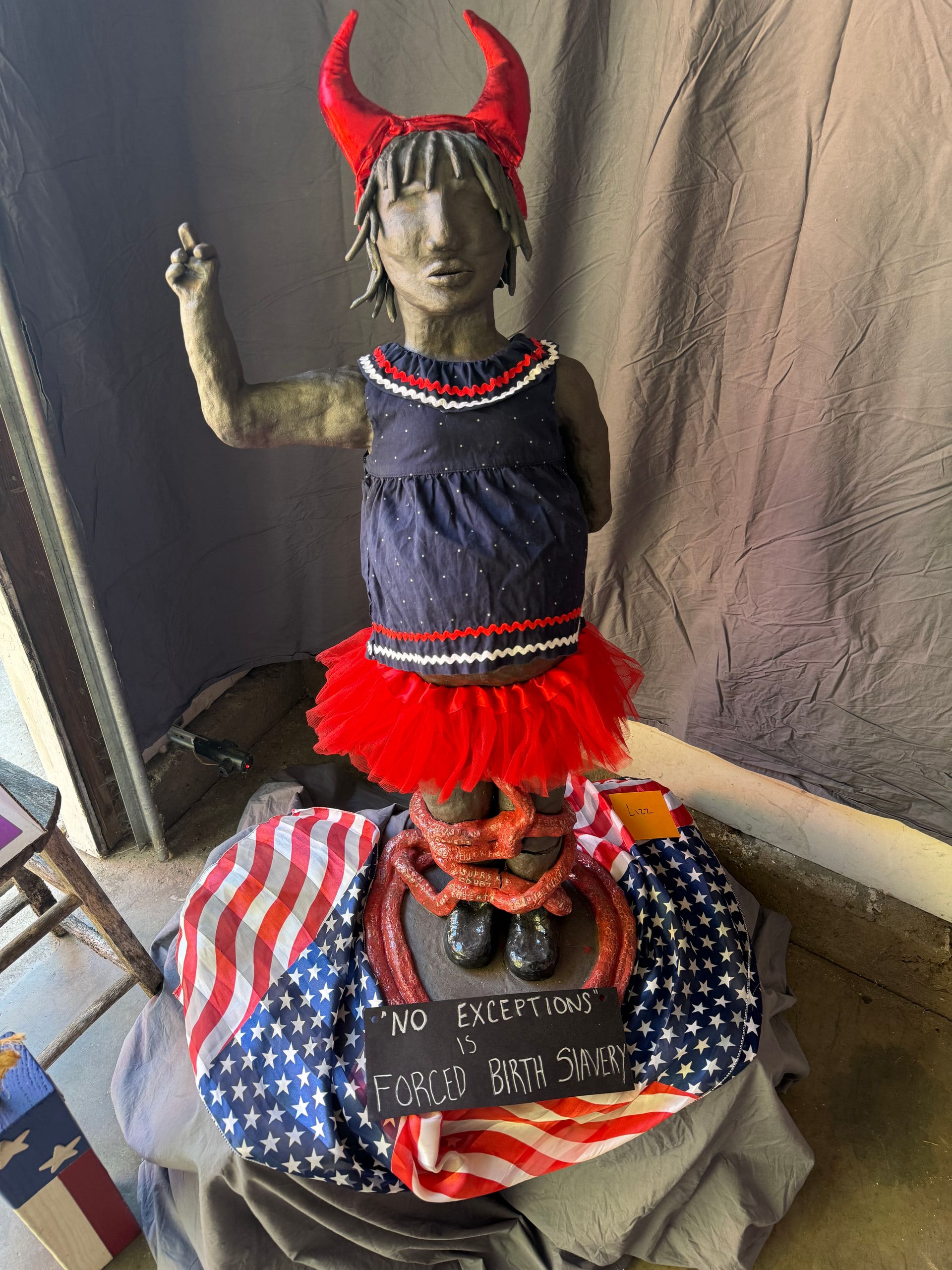 Standing figure with red horns and tutu, a dark dress, and a raised middle finger. Red chain-like forms circle its legs above an American flag and a protest sign.