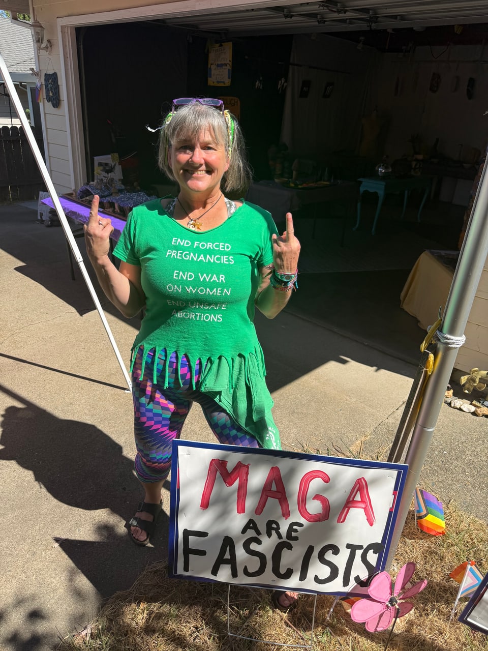 Elizabeth Gallagher outside a studio, wearing a green shirt and raising both middle fingers beside a sign reading “MAGA ARE FASCISTS.”