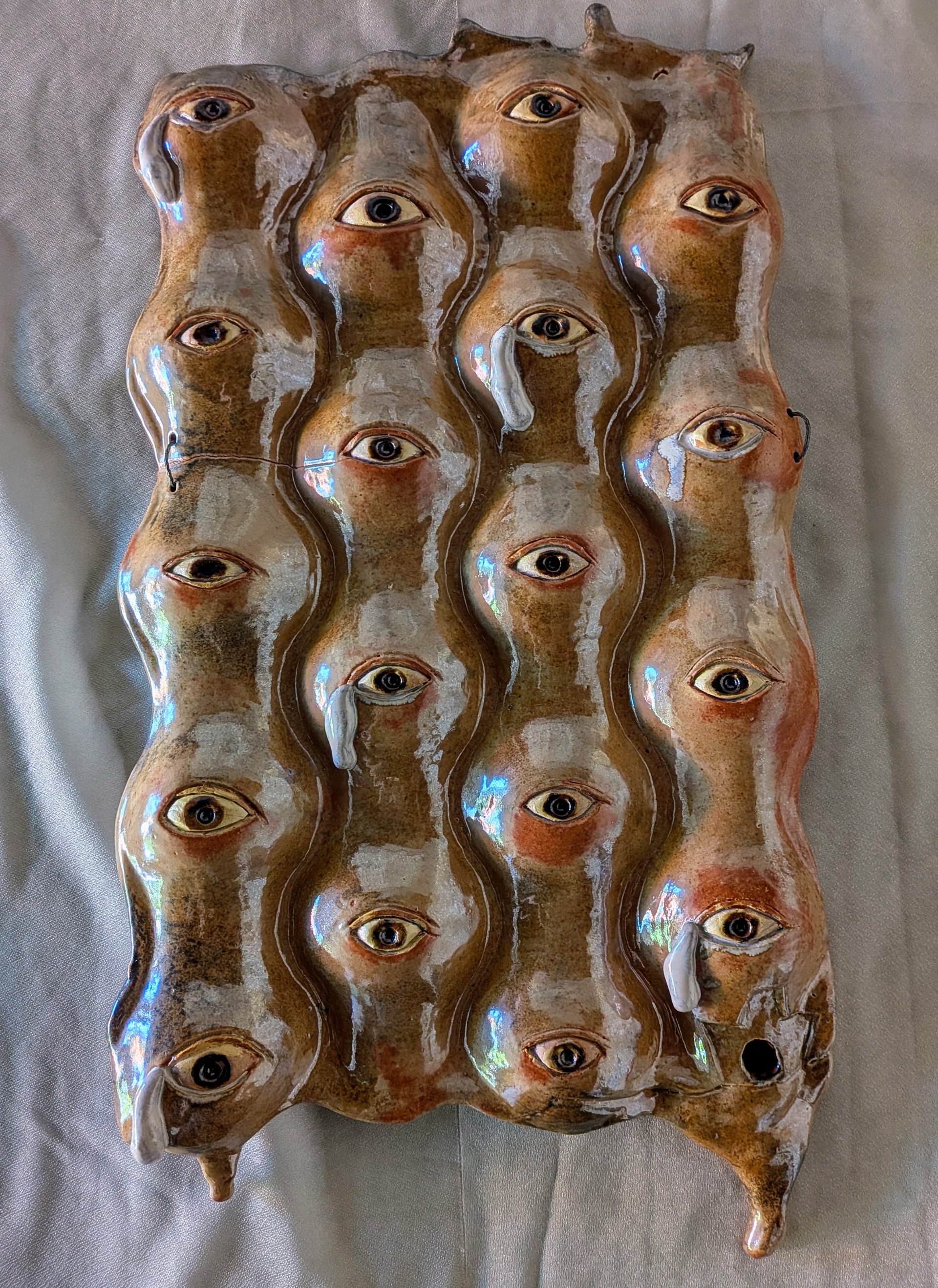A vertically oriented high-fire ceramic sculpture titled Kids in Cages. The piece is a rectangular, undulating slab of brown, tan, cream, and rust-colored glazed clay covered with approximately 20 sculpted human faces. The faces are tightly packed together, with their forms flowing into one another. Each face has a single prominent, realistic-looking eye, creating the impression of many people watching the viewer. Several of the eyes have thick white clay tears running down the faces. The expressions are solemn and weary. The glossy glaze catches blue and white reflections across the surface. The bottom corners of the sculpture extend into pointed, irregular forms, and one face near the lower right has a small dark opening in the clay. The overall effect is of a crowded group of human faces pressed together, crying and looking outward.