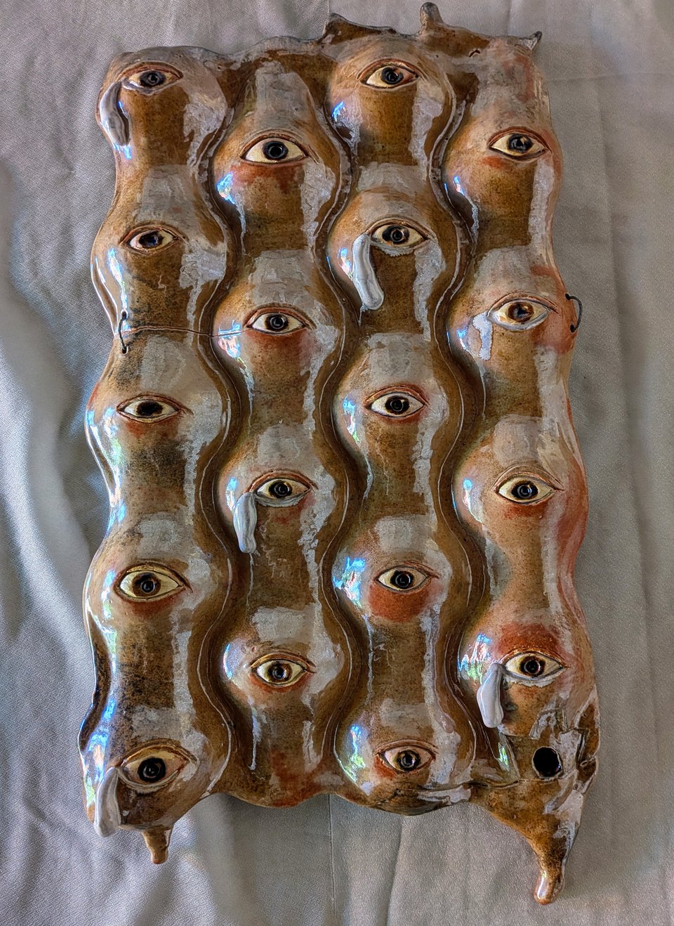 A vertically oriented high-fire ceramic sculpture titled Kids in Cages. The piece is a rectangular, undulating slab of brown, tan, cream, and rust-colored glazed clay covered with approximately 20 sculpted human faces. The faces are tightly packed together, with their forms flowing into one another. Each face has a single prominent, realistic-looking eye, creating the impression of many people watching the viewer. Several of the eyes have thick white clay tears running down the faces. The expressions are solemn and weary. The glossy glaze catches blue and white reflections across the surface. The bottom corners of the sculpture extend into pointed, irregular forms, and one face near the lower right has a small dark opening in the clay. The overall effect is of a crowded group of human faces pressed together, crying and looking outward.